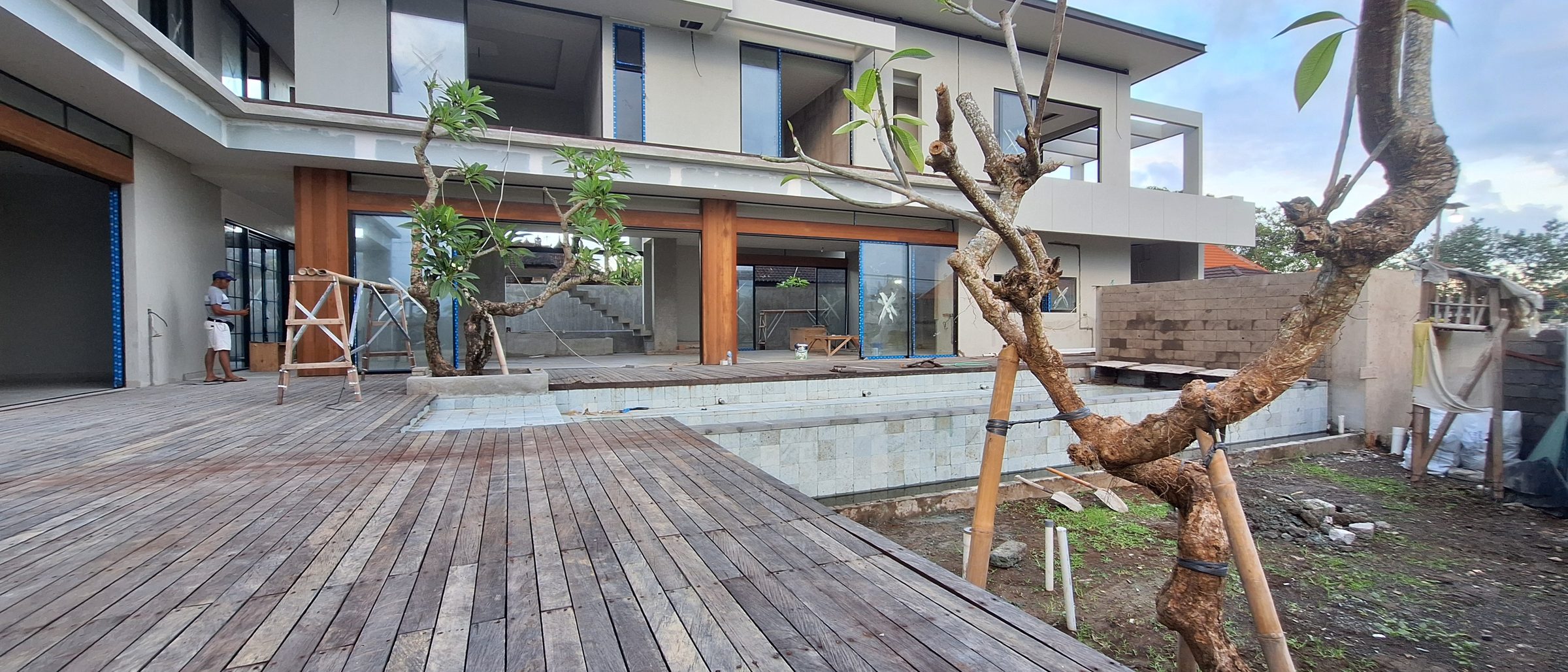 A villa under construction in Bali: a wide timber deck in the foreground, a stone-clad pool wall behind it, bare soil to the right and two frangipani trees held upright by bamboo braces.