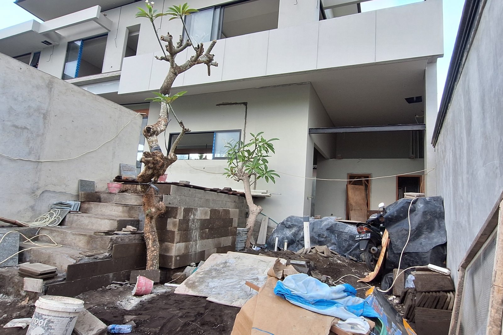 A side yard mid-construction: concrete steps, a low stone retaining wall part-built, stacked materials and two frangipani trees in the ground.